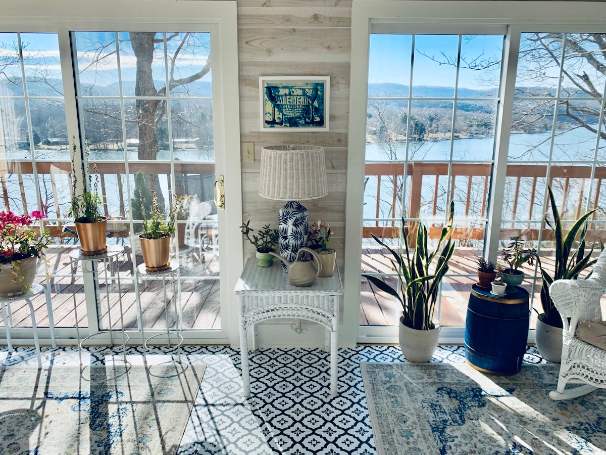 A sunlit sunroom showcases large glass windows framing views of Claytor Lake and the surrounding landscape. Potted plants are arranged on tables, while a decorative lamp sits on a small white table. The room features patterned rugs that enhance the inviting atmosphere.
