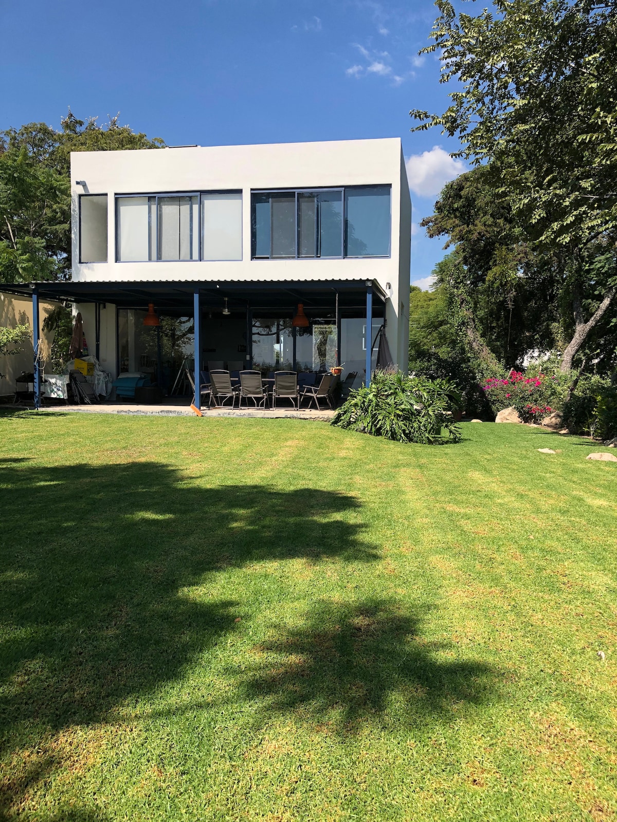 A modern two-story home is set amidst a well-manicured lawn, featuring a covered terrace with multiple chairs. Large windows allow natural light to fill the interior, while surrounding lush greenery enhances the peaceful outdoor atmosphere.