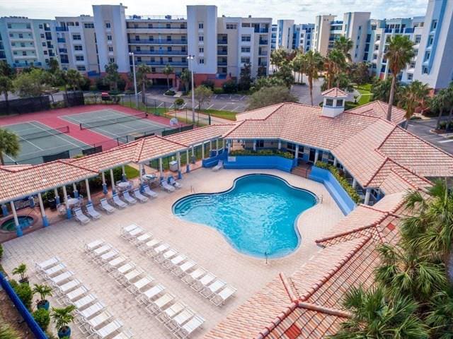 An aerial view captures the resort-style pool surrounded by lounge chairs. The pool area is complemented by a clubhouse featuring a tiled roof. Tennis courts with bright blue surfaces are visible in the background, framed by lush palm trees and well-maintained grounds.