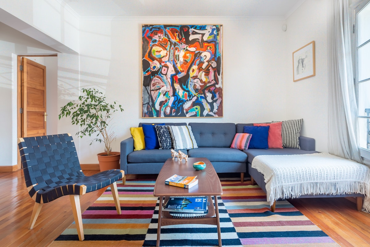 A spacious living area features a modern sectional sofa adorned with colorful cushions. A striped area rug lies beneath a wooden coffee table, which holds decorative items. A large abstract painting hangs on the wall, complemented by a potted plant and bright natural light from the windows.