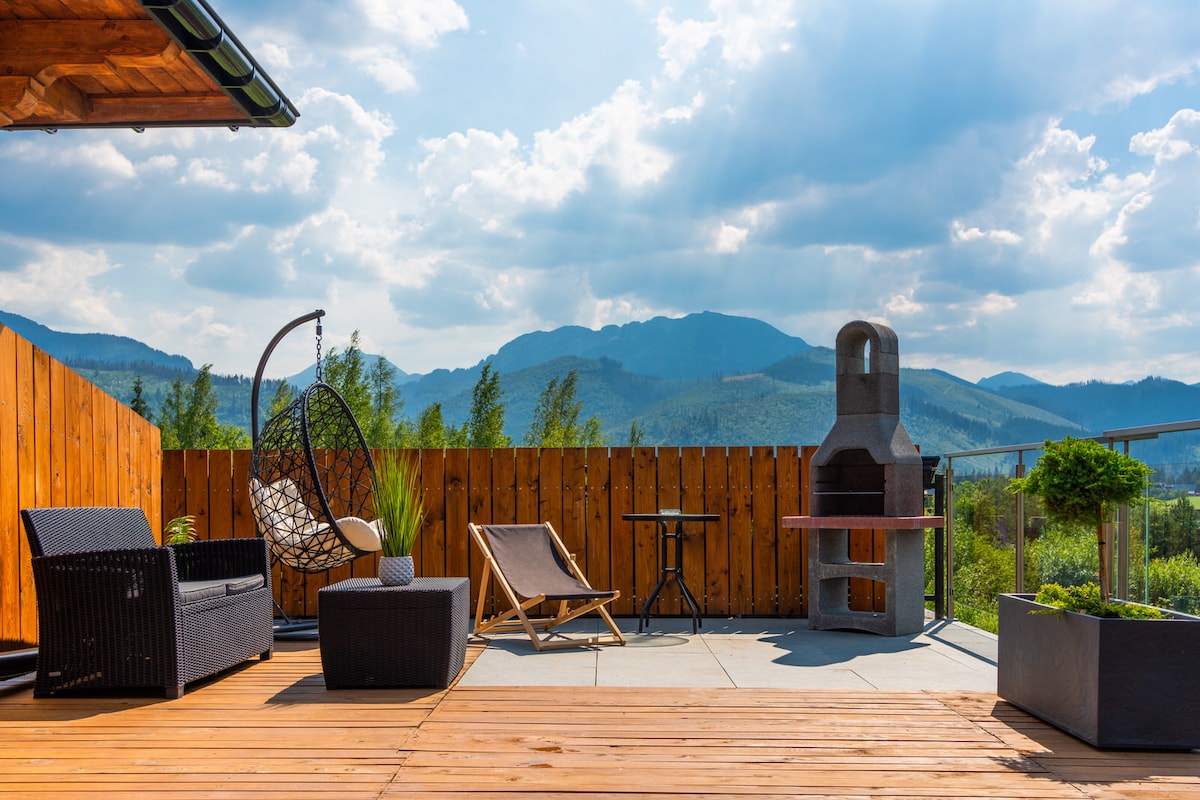 An outdoor terrace features a stylish seating arrangement with a hanging chair and a lounge chair, complemented by a small table. A stone grill adds functionality, while surrounding greenery and a panoramic view of the mountains enhance the tranquil setting.