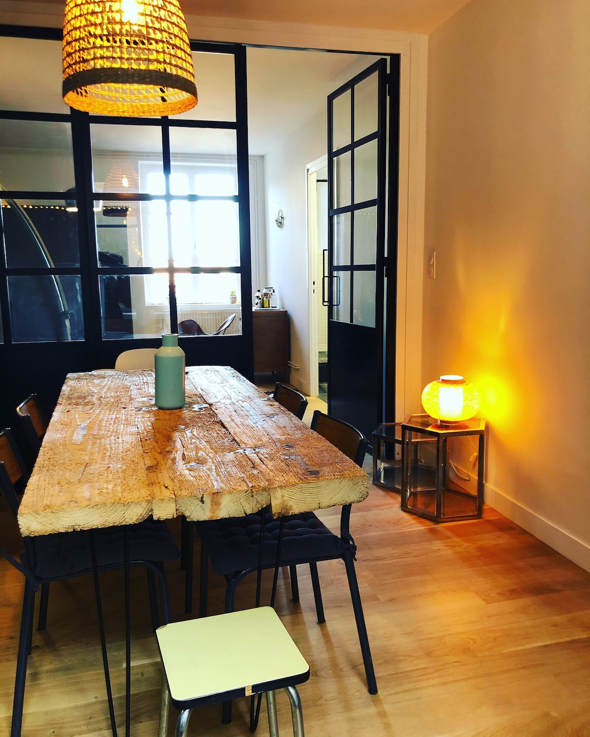 A dining area features a rustic wooden table with simple black chairs arranged around it. A soft glowing lamp provides warm light, while a decorative vase in a calming color rests on the table. Large glass doors lead to an adjoining space.