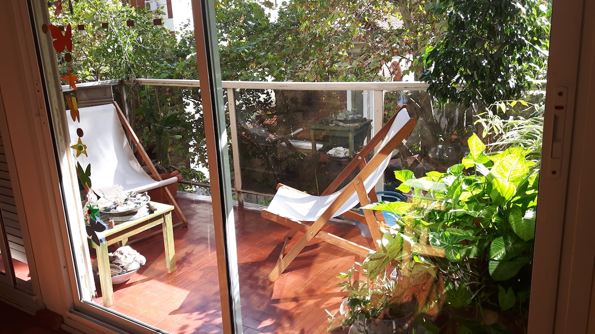 A bright balcony area features a wooden deck with a relaxing lounge chair and a small table. Lush greenery surrounds the space, creating a natural and calming environment. Light filters through the plants, adding to the serene atmosphere.