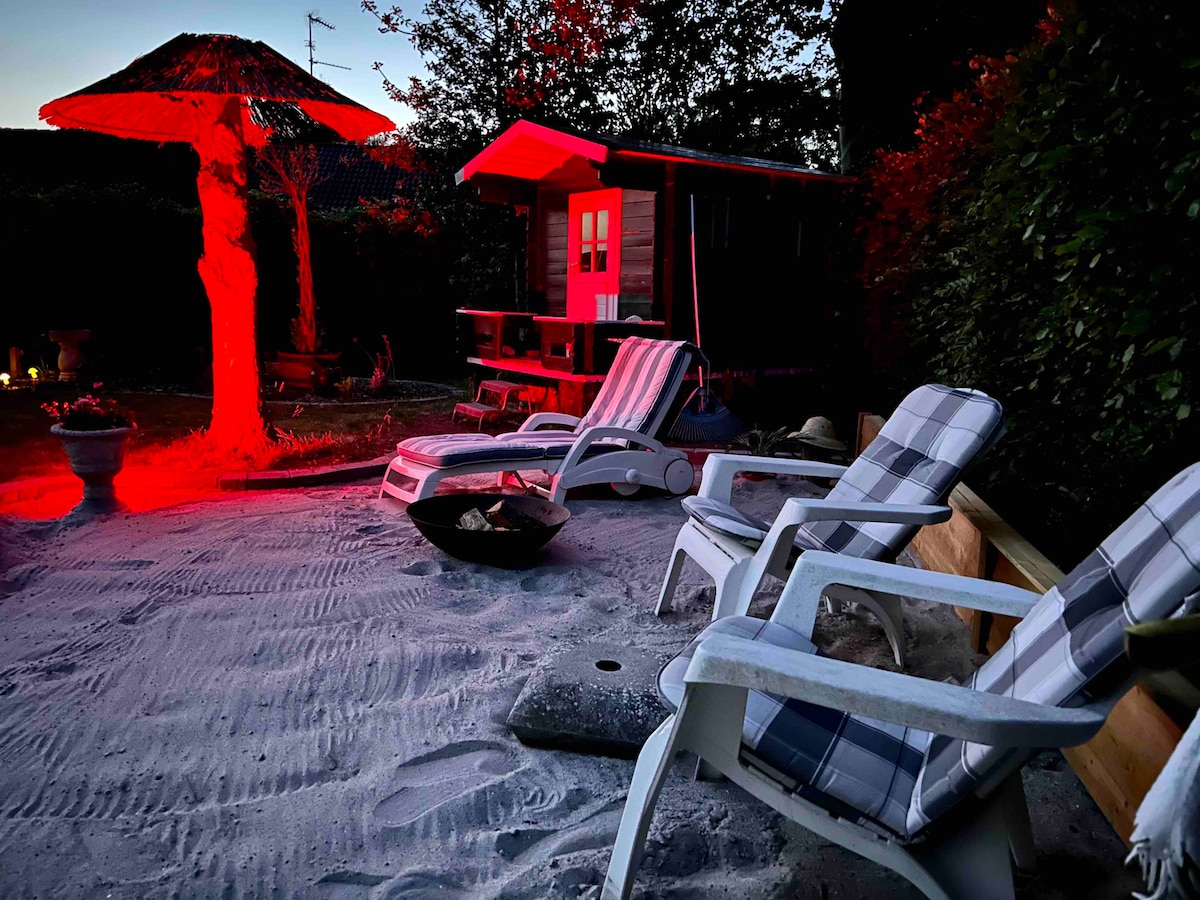 A sandy outdoor area is presented with two white lounge chairs featuring checkered cushions. An umbrella structure provides shade, while a small wooden structure is visible in the background. Soft red lighting illuminates the space, offering a unique ambiance for relaxation.
