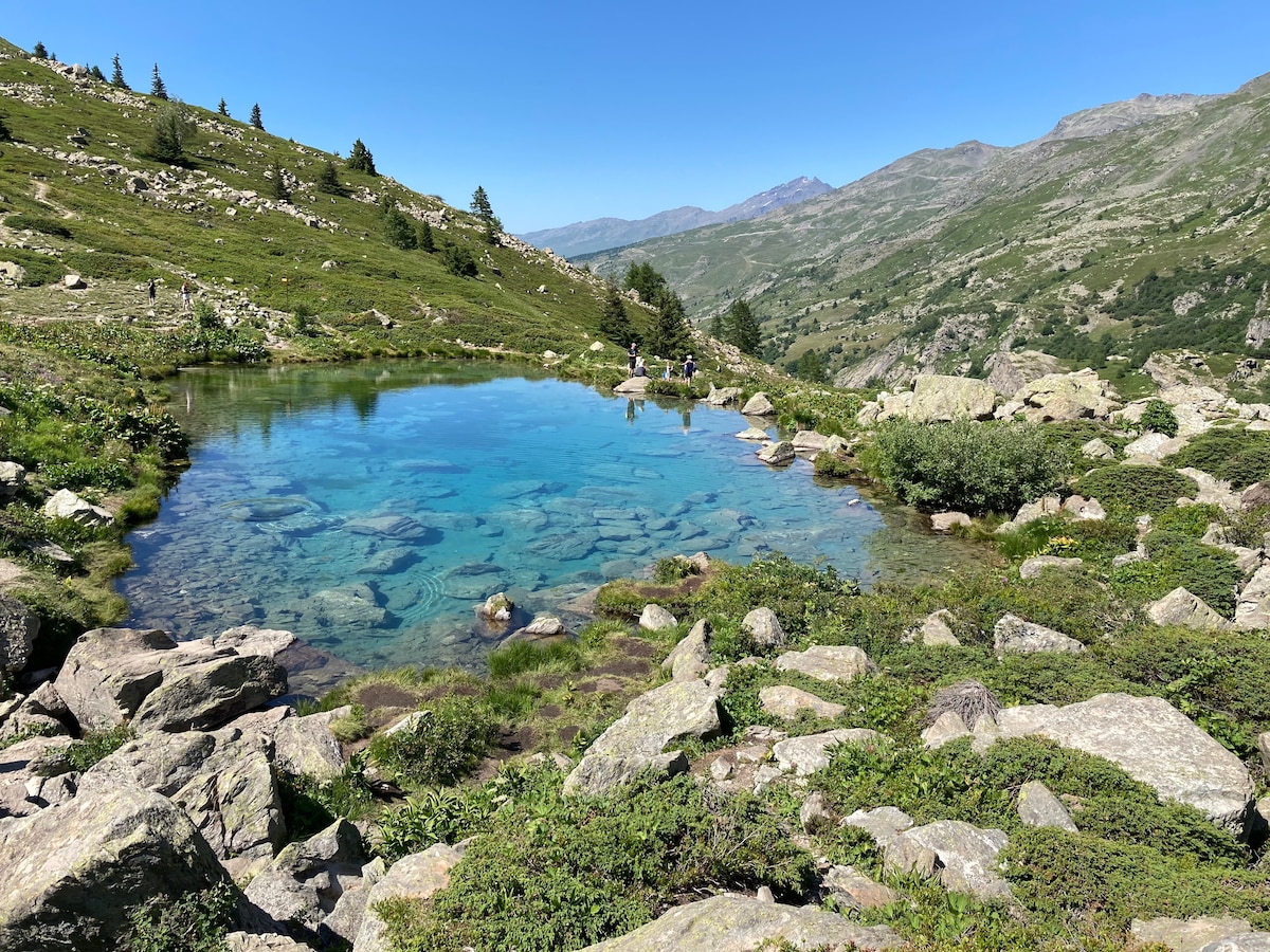 A serene mountain landscape features a clear, reflective pond surrounded by lush greenery and rocky outcrops. Gentle slopes rise in the background, dotted with trees and distant peaks, creating a sense of tranquility in the natural setting.