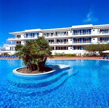 A large swimming pool features a central island of lush palm trees, surrounded by crystal-clear blue water. The modern three-story building with expansive glass balconies stands in the background under a bright blue sky, offering an inviting ambiance.