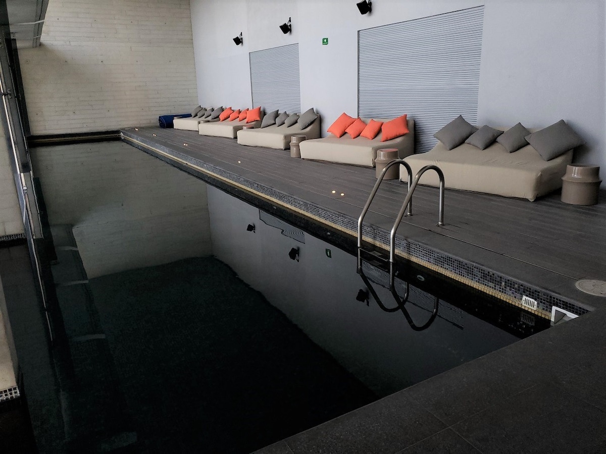 A spacious indoor pool area features a sleek, dark water surface. Plush lounge seating lined along the pool is adorned with cushion accents, and wall-mounted lights provide a subtle glow. The atmosphere is calm, enhanced by the minimalist design of the surrounding space.