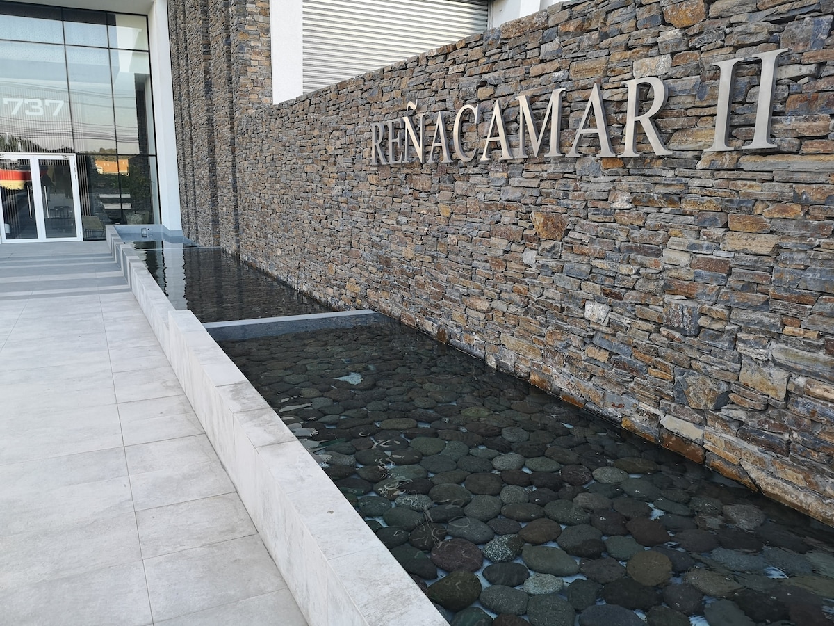 The building entrance features a modern stone wall with 'REÑACAMAR II' elegantly displayed. A shallow water feature lined with smooth stones reflects the surrounding light, enhancing the contemporary aesthetic of the space. Paved pathways lead toward the entrance, providing a welcoming approach.