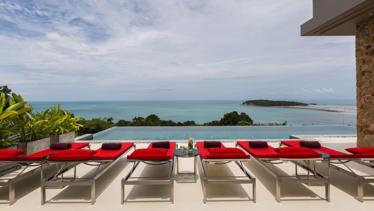 A serene terrace is featured, lined with red lounge chairs overlooking a sparkling infinity pool. The expansive view reveals the tranquil ocean and lush greenery, enhancing the space's connection with nature.