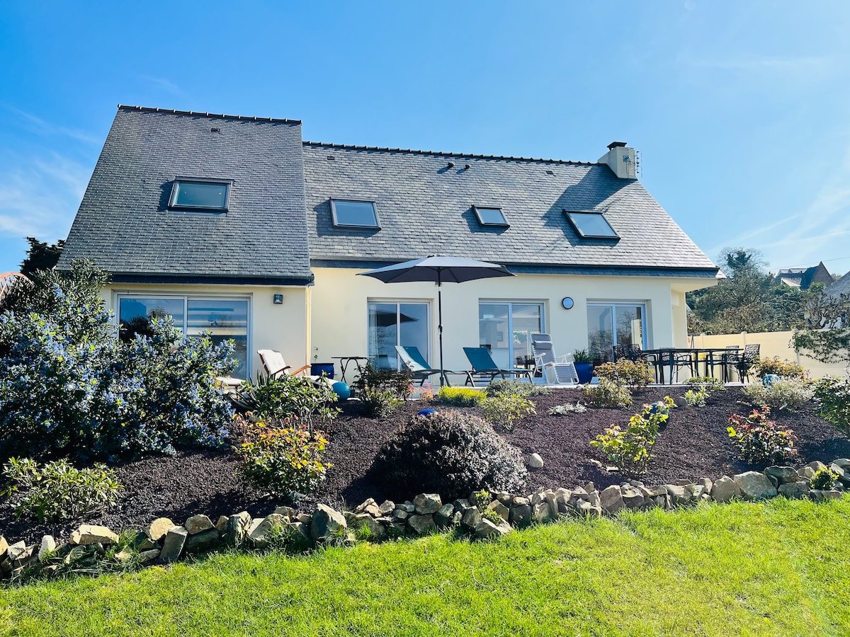 The exterior of the house is visible, featuring a sloped roof and large windows that invite natural light. A garden with various plants frames the home, while a patio area with seating and an umbrella is set up for outdoor relaxation. A clear blue sky enhances the setting.