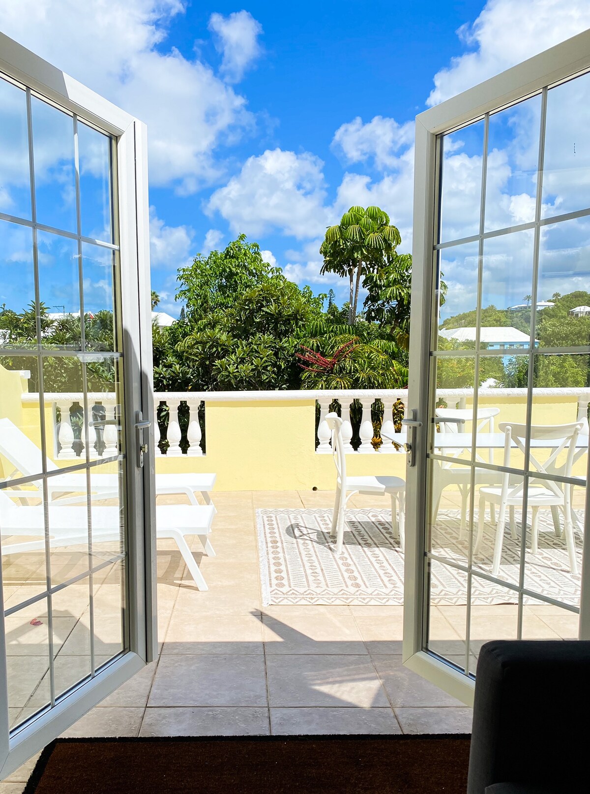 Double glass doors open to a sunny patio featuring white loungers and a dining table surrounded by greenery. A patterned outdoor rug sits on tiled flooring, complementing the bright blue sky with fluffy clouds in the background.