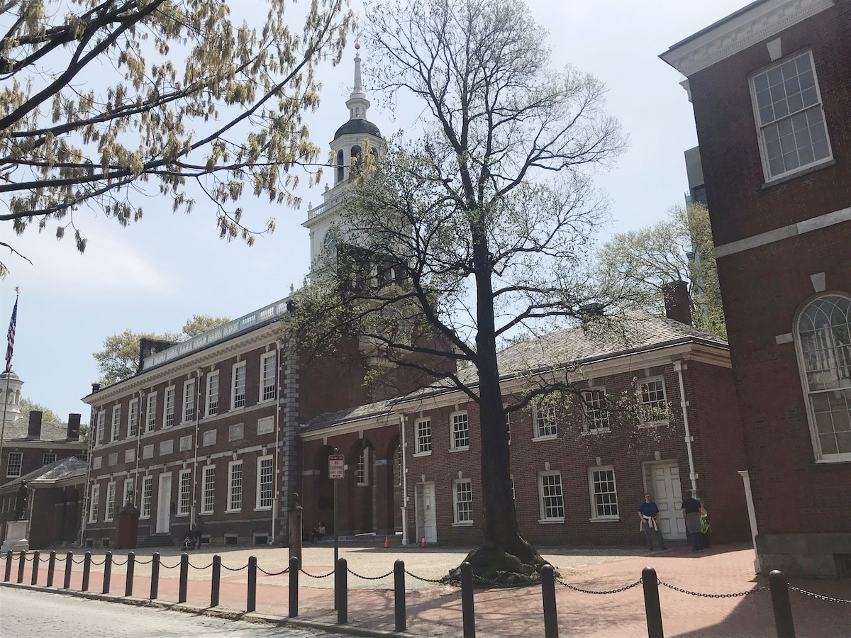 A historic brick building stands prominently in the foreground, featuring a classic clock tower. An expansive tree can be seen nearby, with light foliage. A cobblestone path borders the site, hinting at its historical significance and inviting exploration.