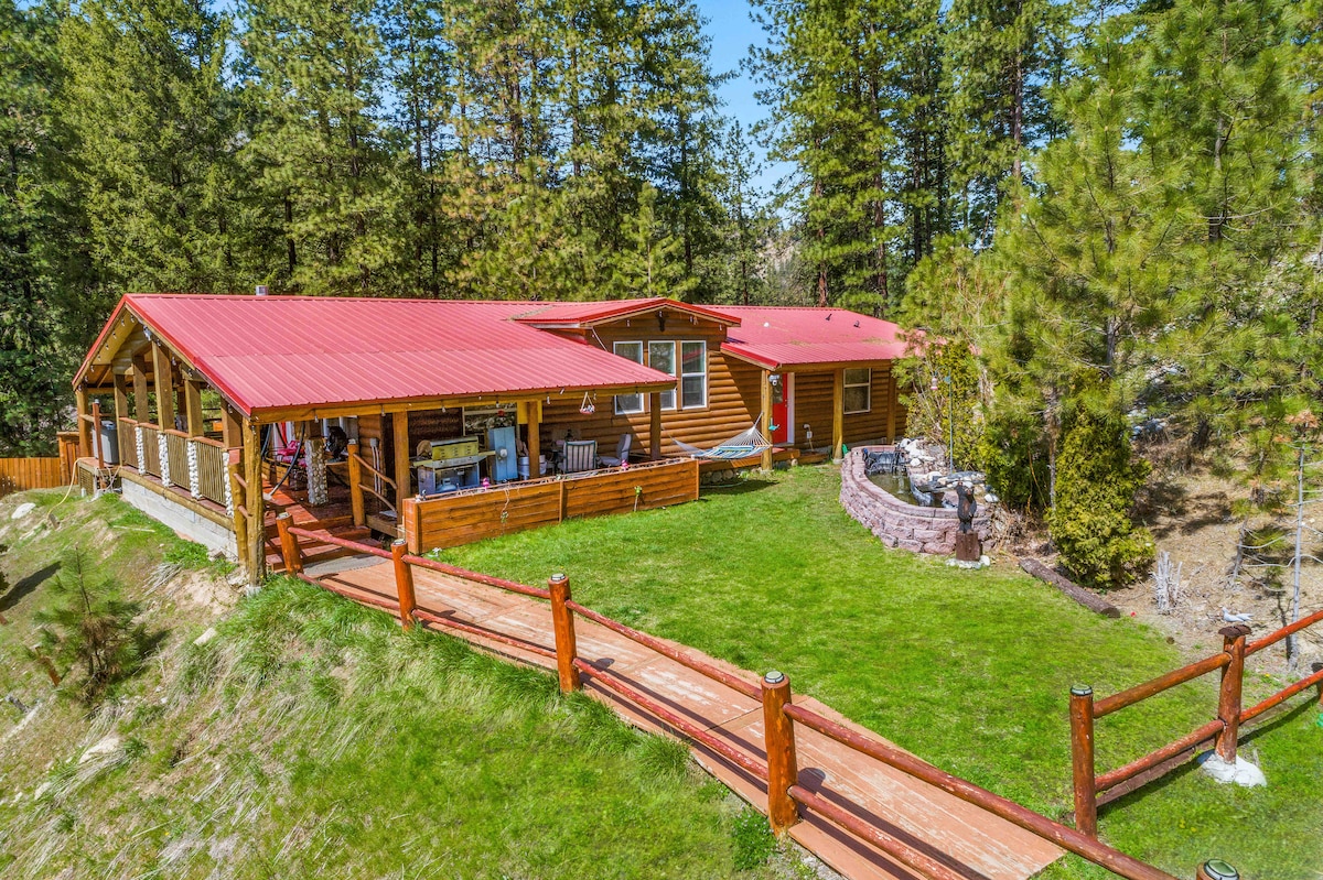The cabin features a red roof and is surrounded by lush greenery. A spacious porch extends along the front, furnished for outdoor relaxation. A well-maintained lawn leads to a seating area with a fire pit, complemented by natural landscaping that enhances the serene environment.