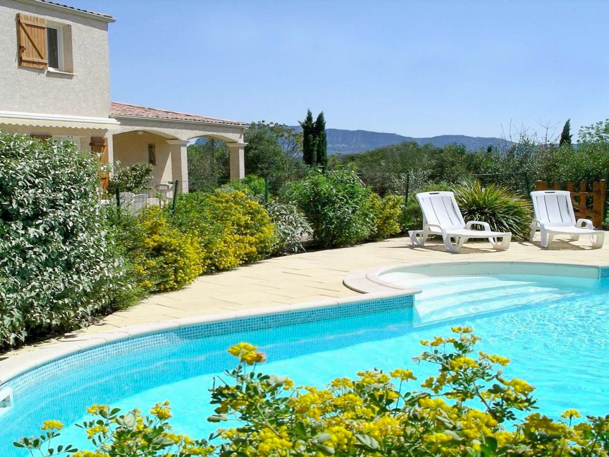 A private pool is surrounded by landscaped gardens featuring vibrant yellow flowers. Sun loungers are positioned next to the pool, providing a relaxing space. The backdrop includes lush greenery and hills, creating a serene outdoor atmosphere.