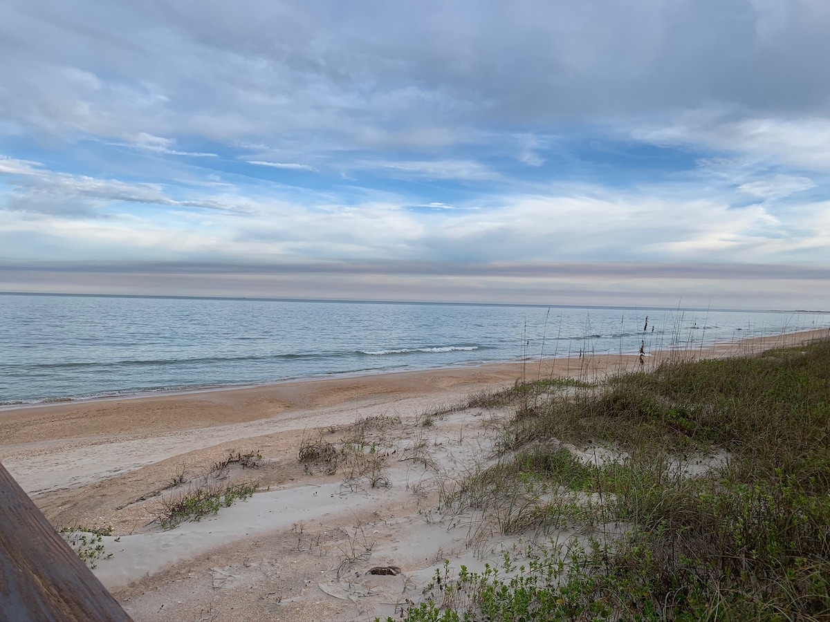 The tranquil beachfront is depicted under a cloudy sky, with soft waves lapping at the shore. Sandy dunes covered with sparse greenery lead towards the water's edge, creating a serene coastal atmosphere. A distant figure strolls along the beach, enjoying the expansive ocean view.
