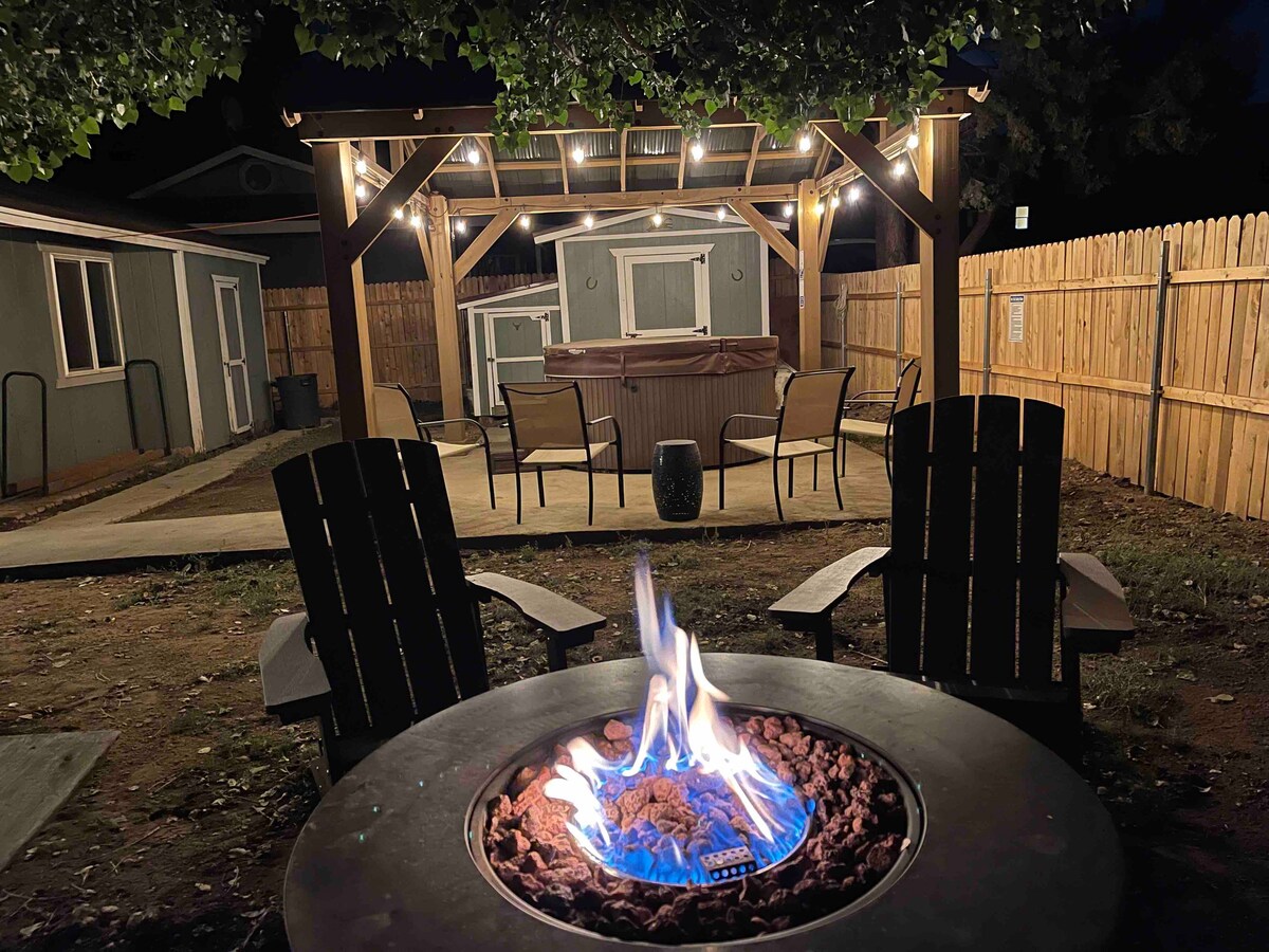 A well-lit outdoor space features a wooden pergola above a hot tub surrounded by a paved patio. Two adirondack chairs are positioned beside a gas firepit, where flames dance above decorative rocks. A wooden fence encloses this inviting area, creating a private setting.