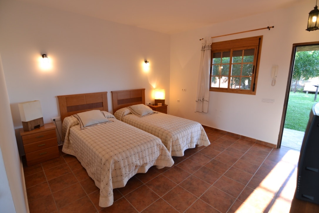 A spacious bedroom is showcased with two separate beds, each dressed in light bedding. Natural light enters through a window next to a wooden door that leads outside. Soft wall sconces provide additional illumination, and wooden nightstands are positioned beside the beds.