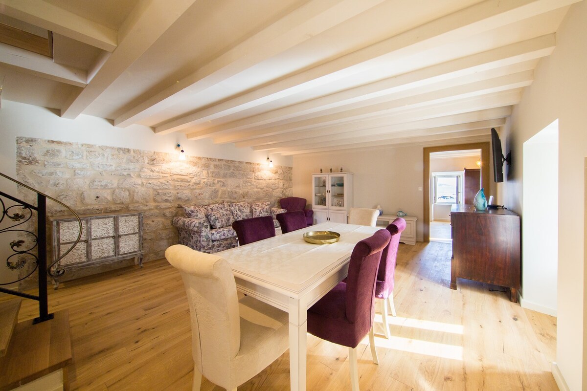 A spacious living area features exposed wooden beams and pale wooden flooring. A large table surrounded by upholstered chairs is positioned in the center, while a comfortable seating arrangement is situated along the stone wall. Natural light illuminates the room from large windows.