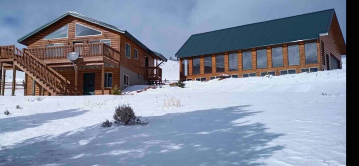 Two rustic wooden structures are positioned amid a snowy landscape. One features a balcony and a steep staircase, while the other showcases large windows allowing natural light inside. The untouched snow blankets the ground, creating a serene winter atmosphere.