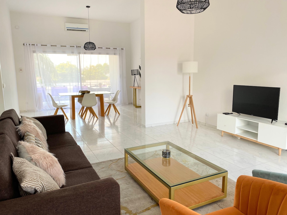 A bright living area features a comfortable dark sofa and an orange armchair, arranged around a stylish coffee table. Large glass doors provide natural light and access to a terrace. A flat-screen TV is positioned on a low white cabinet, complemented by modern light fixtures.