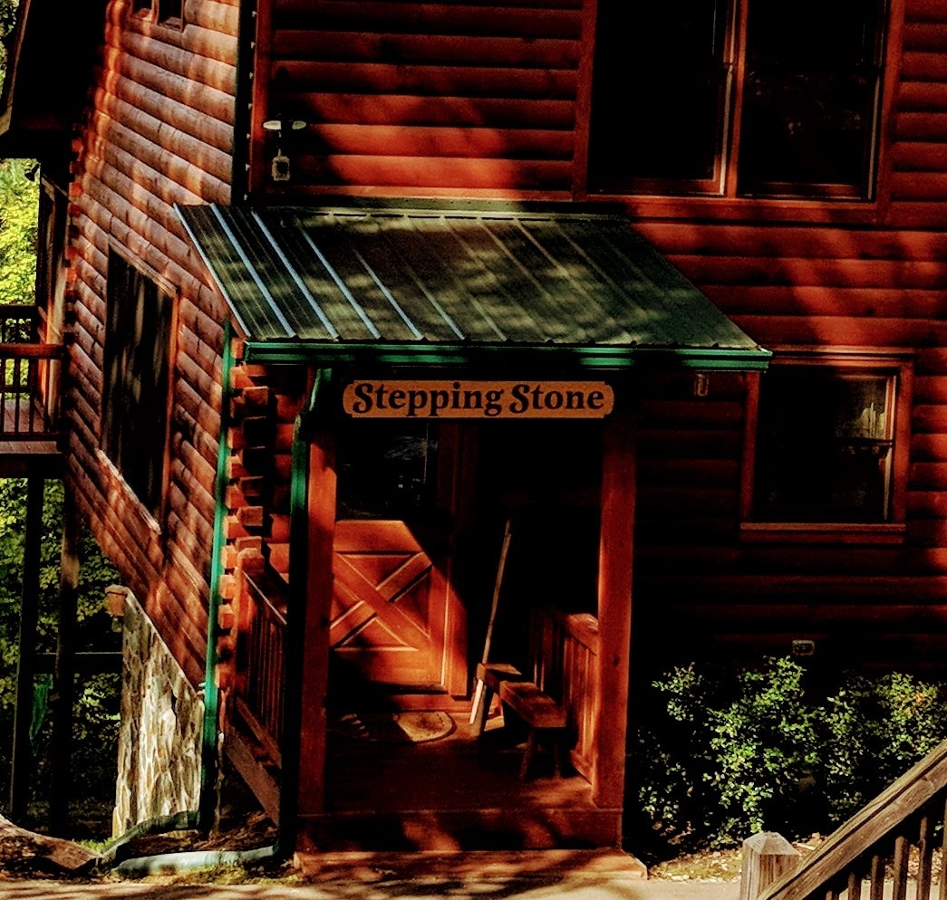 The cabin's entrance showcases a welcoming wooden porch with a sloped roof. The 'Stepping Stone' sign is prominently displayed above the door, flanked by large, well-placed windows. Natural light filters through, illuminating the warm wooden tones of the log exterior.