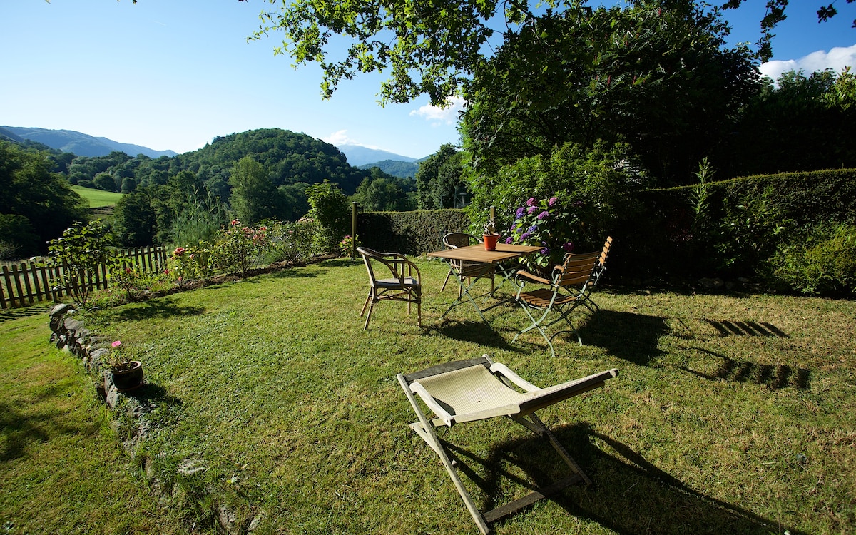 A neatly maintained garden area features a table and chairs, surrounded by greenery. The landscape offers views of distant hills under a clear blue sky. Bright flowers add color to the natural setting, creating a serene environment for relaxation.