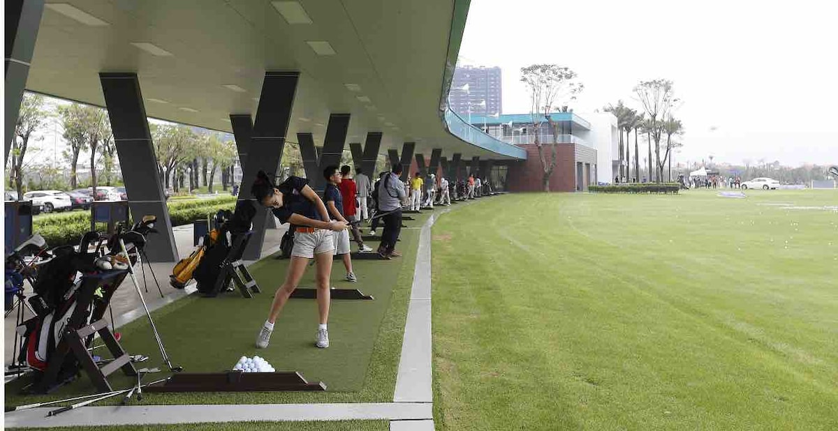 A golf driving range is displayed with multiple practice bays covered in artificial turf. Several golfers are seen preparing to swing their clubs, with golf bags positioned next to them. A spacious area with greenery is visible in the background, creating a serene environment.