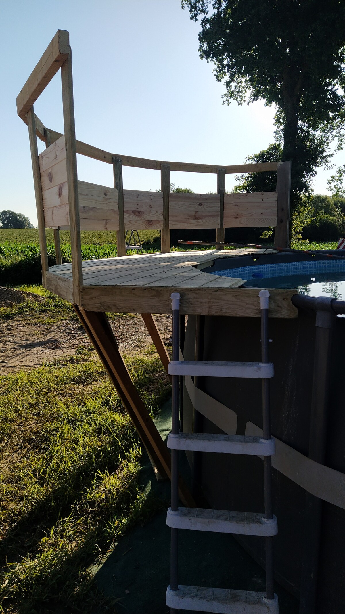 A wooden deck is constructed around an above-ground pool, providing a space for sitting or relaxing. A ladder leads to the pool, surrounded by grassy areas and crops in the background. The clear sky enhances the sense of openness in the outdoor environment.