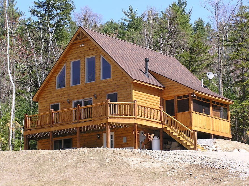 A log-sided chalet style home is featured with a spacious deck surrounding the front. Large windows provide natural light and views of the surrounding pine trees. The exterior showcases a blend of wooden textures and earthy tones, while a staircase leads to the entrance.