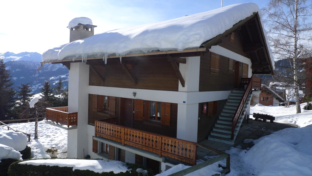 The chalet-style building features a wooden exterior with a sloped roof, covered in snow. Multiple balconies are visible, and a staircase leads to the entrance. Surrounding the structure, snow blankets the ground, creating a serene winter landscape.