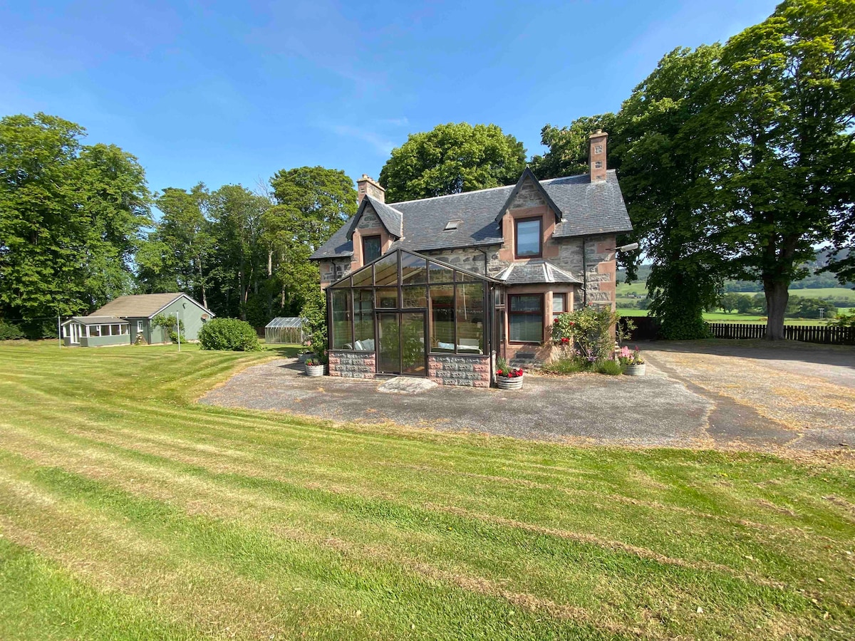 A charming two-story house is surrounded by lush greenery, featuring a large glass conservatory at the front. The manicured lawn and gravel driveway lead up to the entrance, offering an inviting approach. A separate outbuilding can be seen in the background.