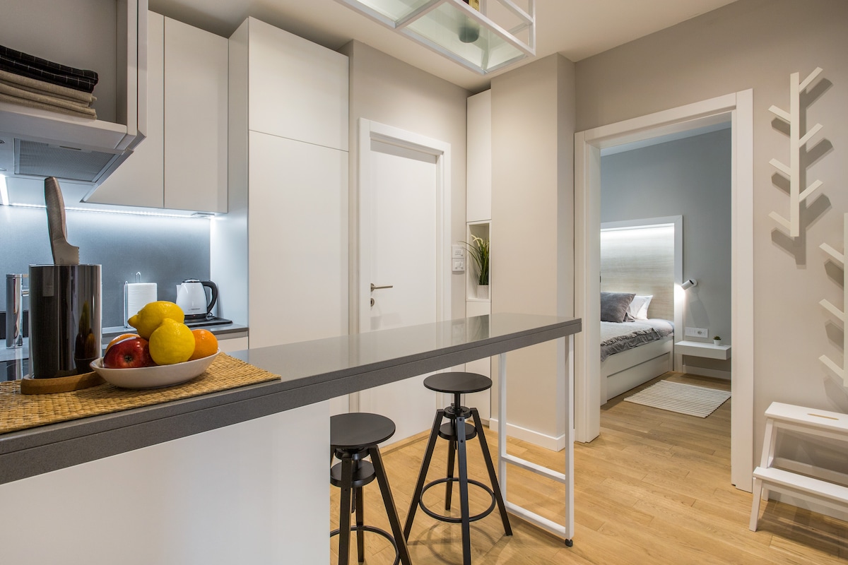 A modern kitchen area is shown, featuring a sleek grey island with two black stools. A variety of fresh fruit is arranged on a woven mat. The light wood flooring complements the minimalist cabinetry and leads into a softly lit living area visible through an open door.