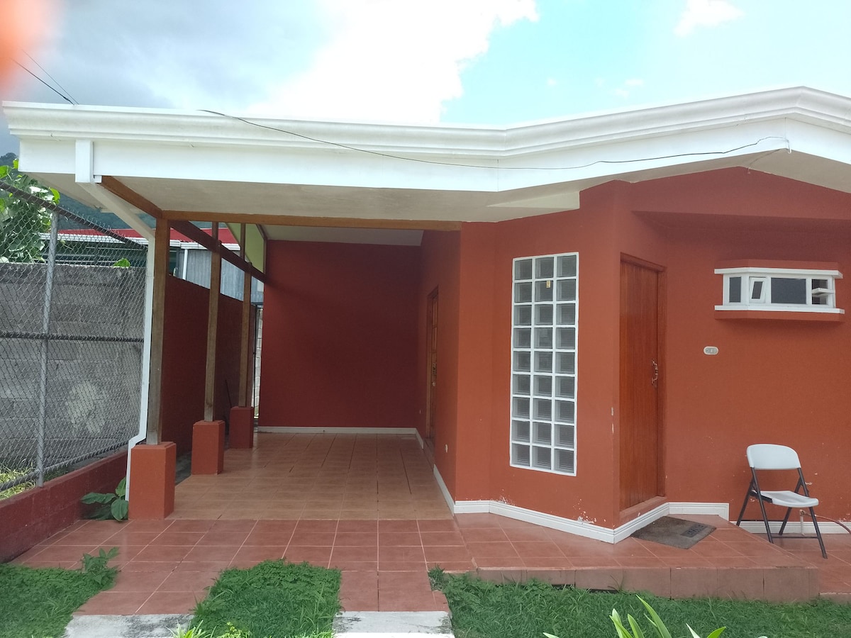 The exterior of the house features a vibrant orange facade and a covered porch area. A small white chair is visible on the porch. The surrounding grass is well-maintained, and the entrance is marked by a simple door and window designs.