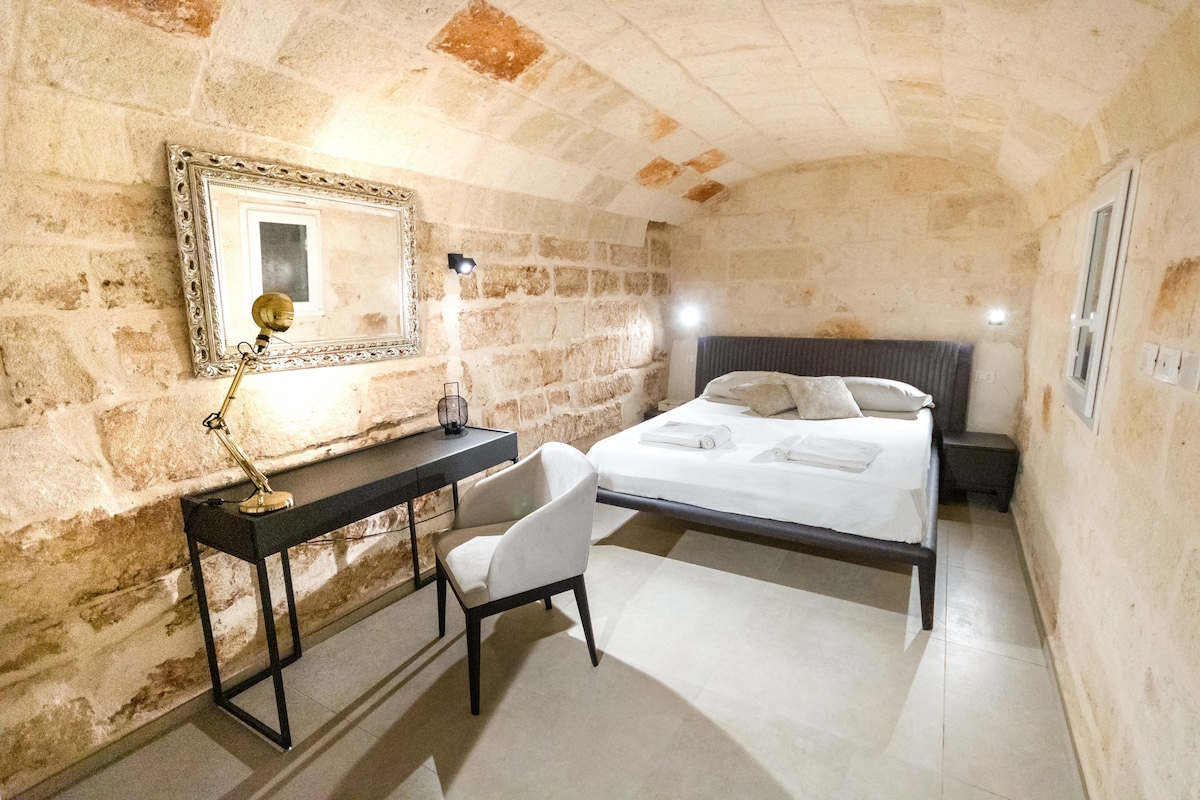 A spacious bedroom features stone walls and a vaulted ceiling, creating a rustic ambiance. A large bed is flanked by two bedside lamps. A sleek desk and chair are positioned in one corner, providing a functional area for work or relaxation.