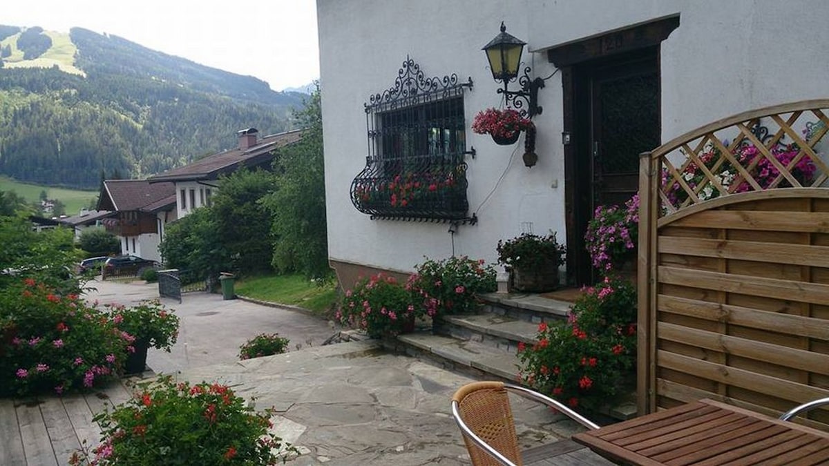 The entrance to the building is framed by vibrant flower arrangements in planters. A wooden fence provides a division, while a small outdoor seating area features a round table and chairs. Lush greenery and hills are visible in the background.