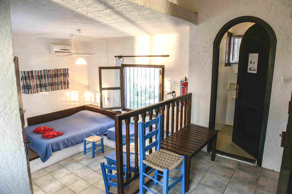 A spacious room features a built-in double bed with red pillows and a single bed, complemented by blue chairs around a wooden table. Natural light filters through a window, illuminating the tiled floor and enhancing the airy atmosphere.