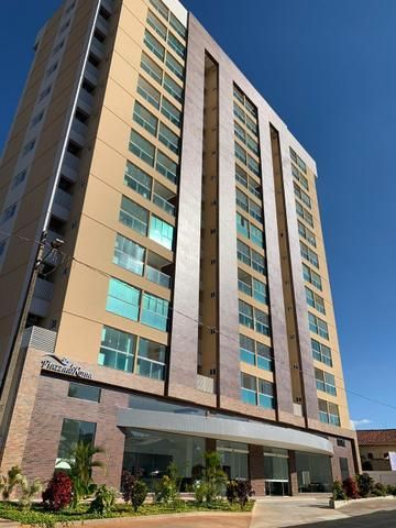 A modern multi-story building is presented with large glass windows, allowing natural light to fill the interior spaces. The facade features a combination of light-colored stone and brick materials, framed by landscaped greenery in the foreground.