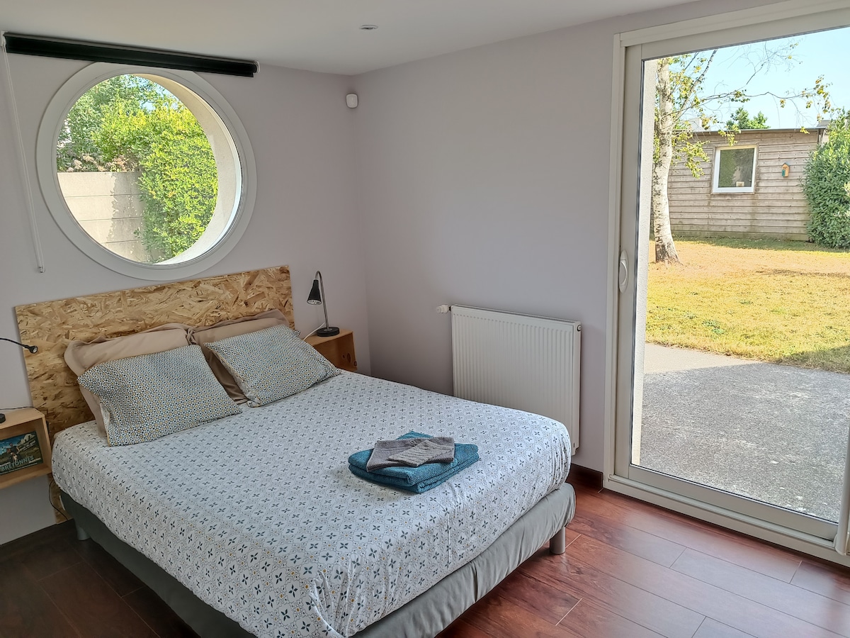 A spacious sleeping area features a comfortable double bed, adorned with soft linens and neatly arranged towels. A circular window allows natural light to illuminate the room, while a large glass door opens to a grassy outdoor area.