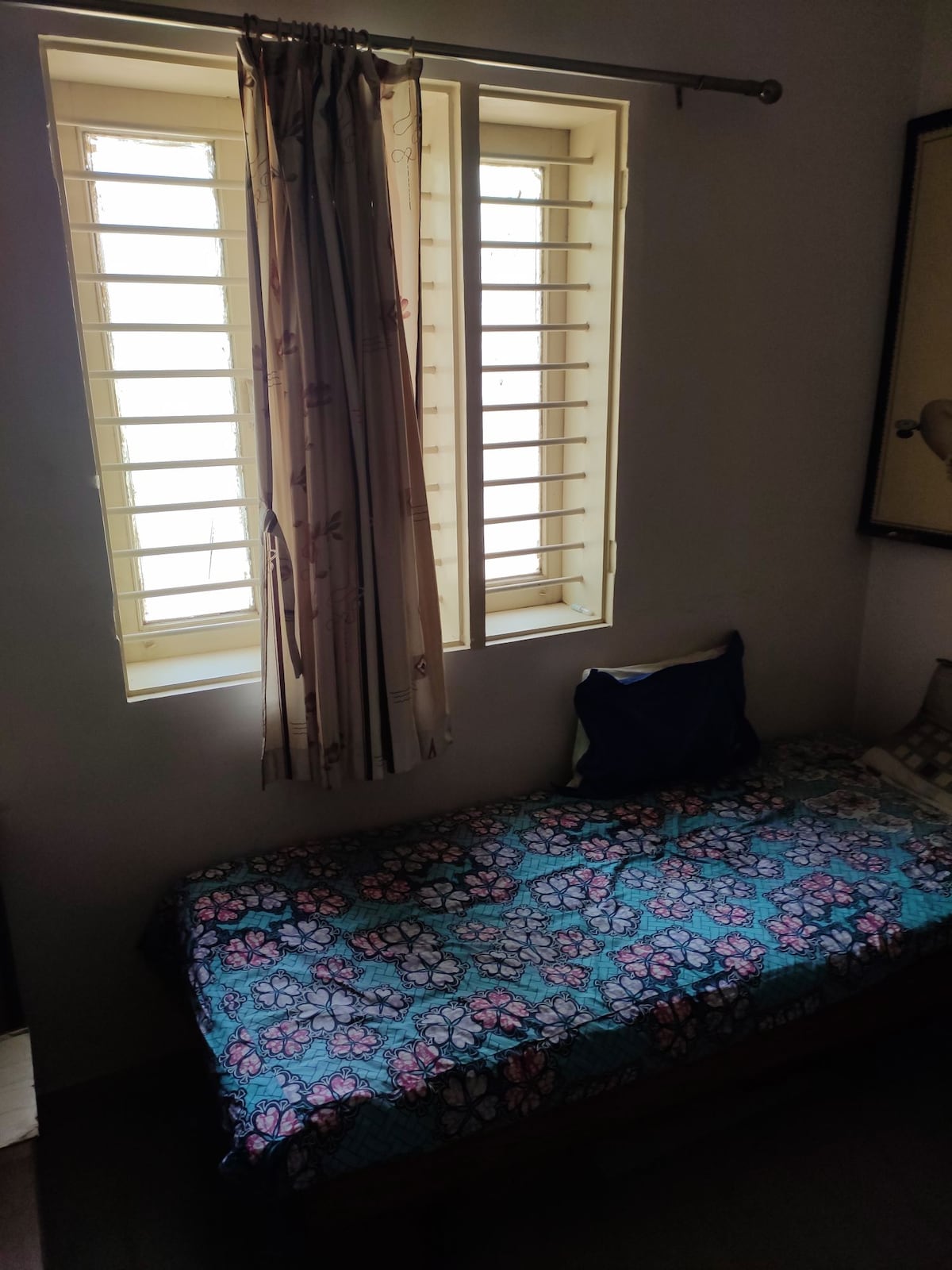 A cozy sleeping area contains a single bed adorned with a colorful floral bedspread. Light enters through two windows with wooden slats, casting soft shadows. A pillow rests against the wall, while a framed picture adds a decorative touch.