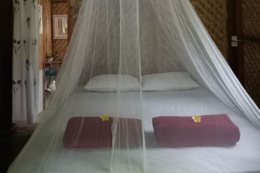 A spacious bed is draped with a sheer white mosquito net, creating a serene sleeping environment. Two towels, each adorned with a yellow flower, are neatly placed at the foot of the bed. The background features natural bamboo walls, enhancing the room's earthy aesthetic.