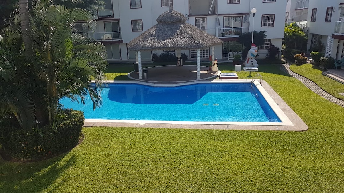 A well-maintained pool area is surrounded by a vibrant green lawn and tropical plants. A palapa-style gazebo provides shaded seating, while a few lounge chairs are positioned near the sparkling blue water of the pool, inviting relaxation and leisure.