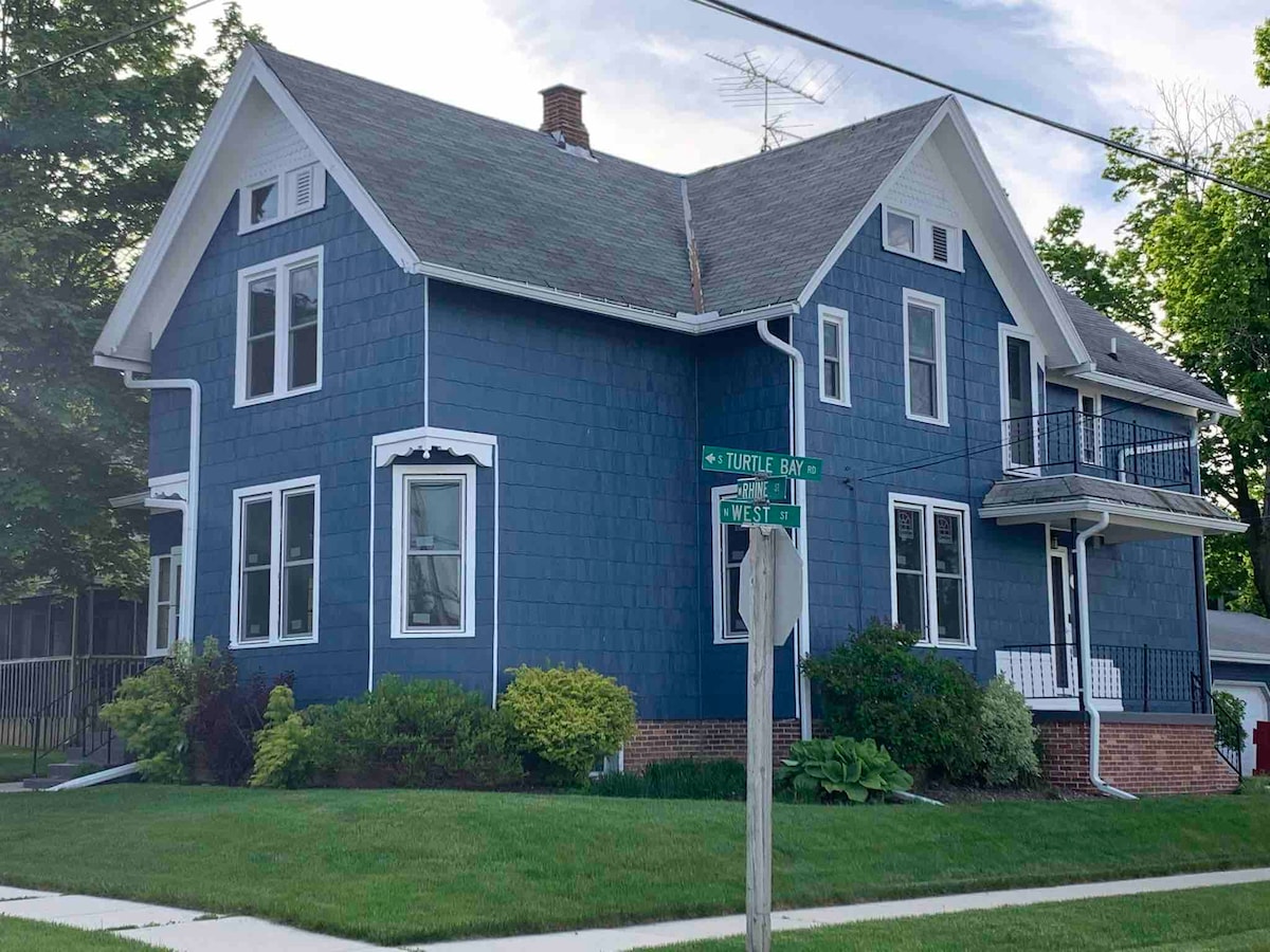 A charming blue duplex is prominently positioned at the corner of two streets. Lush greenery surrounds the building, and a well-maintained lawn is visible in front. The structure features multiple windows and access to a small balcony on the upper level.