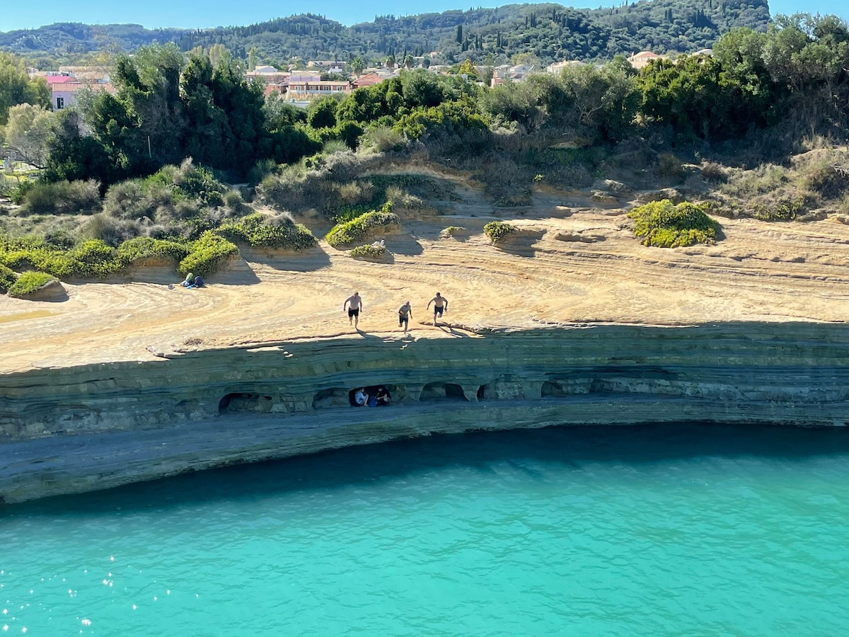 A rugged shoreline is depicted with smooth, layered rock formations alongside clear turquoise water. A group of individuals can be seen exploring the area, while lush greenery and distant hills provide a scenic backdrop.