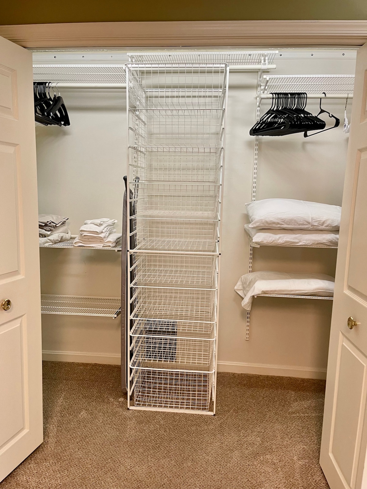 A well-organized closet with open shelving and a white wire storage unit is shown. Neatly folded towels are stacked on shelves, while hangers are provided for clothing. The space is illuminated and carpeted, creating a clean and tidy atmosphere.