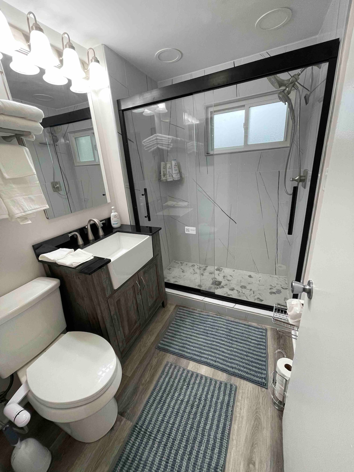 The bathroom features a modern design with a glass shower enclosure and a sleek vanity with a single sink. Towels are neatly arranged on a rack, while a small rug lies on the floor. Natural light enters through a window, enhancing the space's brightness.
