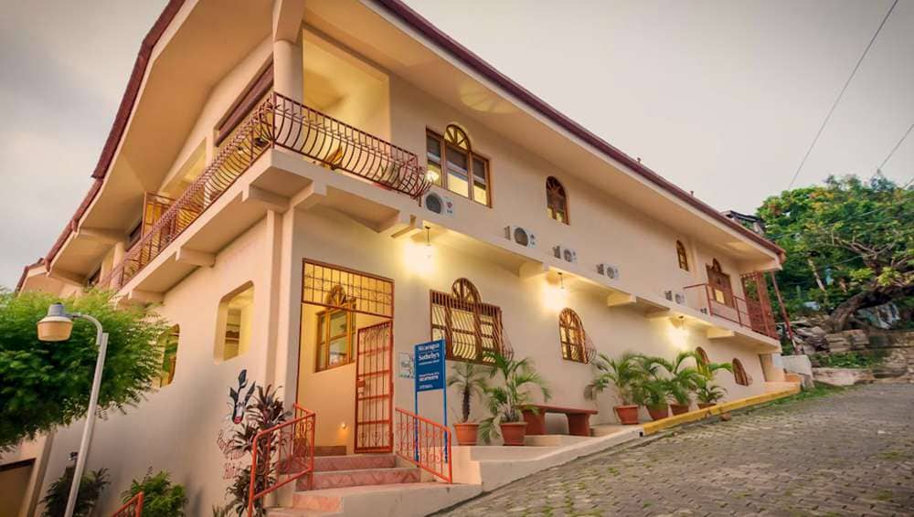 The exterior of the condo is showcased, featuring a two-story design with arched windows and a balcony. Potted plants line the entrance, and warm lighting accentuates the building's inviting façade. A paved area leads up to the structure, with additional greenery in the background.