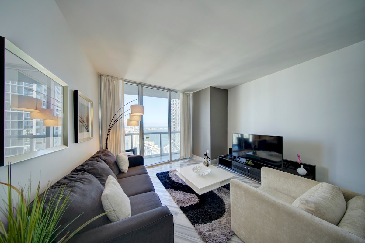 A modern living area is visible, featuring a plush sectional sofa and a comfortable armchair. A sleek coffee table sits at the center, while a large flat-screen TV is positioned against the wall. Floor-to-ceiling windows allow natural light to illuminate the space, offering views of the city.
