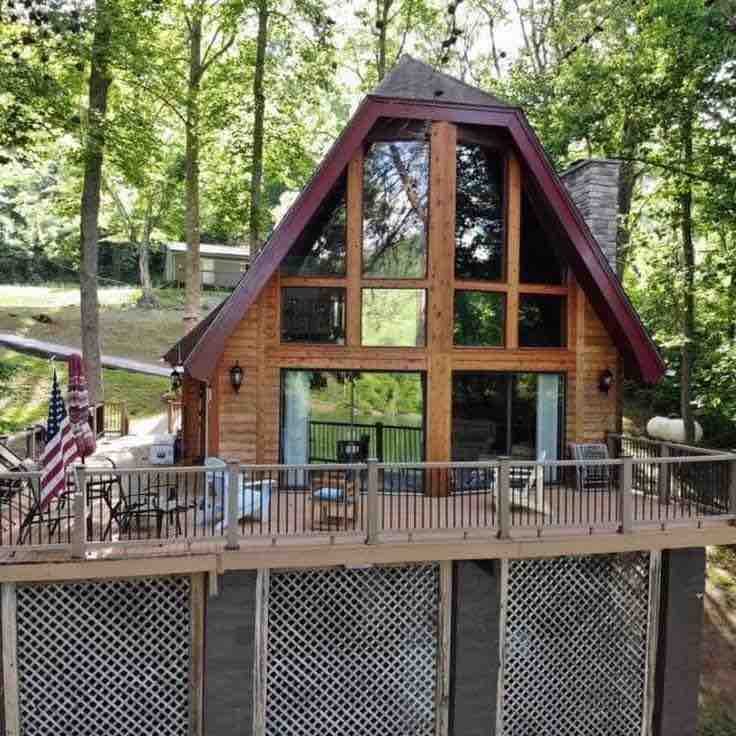 A charming A-frame cabin is positioned among tall trees, featuring large windows that provide ample natural light. A spacious deck surrounds the cabin, equipped with seating and a railing. The exterior showcases wooden finishes, complemented by green landscaping.