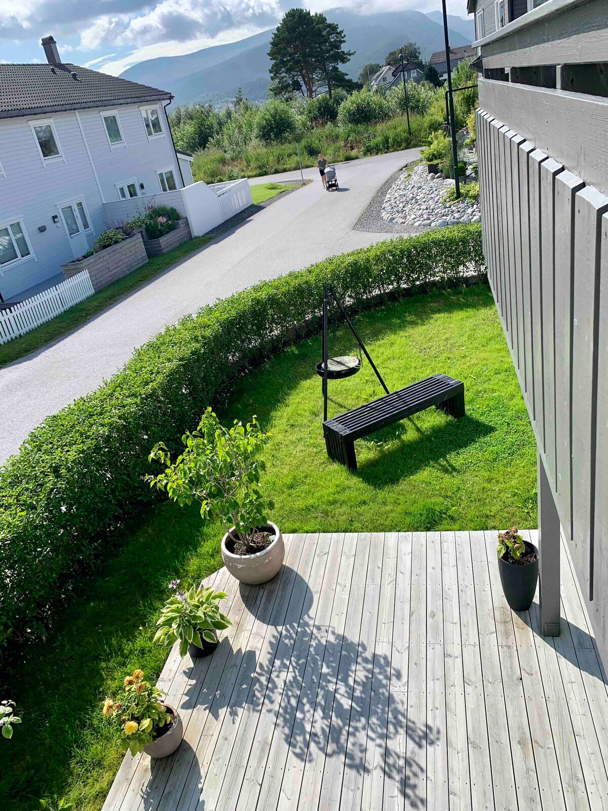 A view from a balcony showcases a well-maintained garden area with a green lawn and a swing seat. Potted plants are visible along the decking. A quiet street is seen in the background, bordered by neat hedges and residential homes.