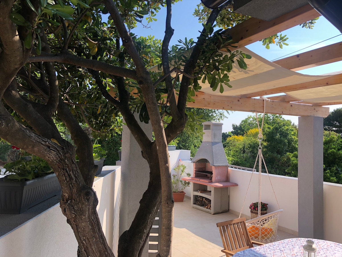 An outdoor terrace features a canopy providing shade, surrounded by greenery. A light-colored outdoor dining set is positioned nearby. A built-in barbecue adds to the space, while a hanging chair offers a cozy spot to relax and enjoy the surrounding nature.